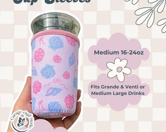 Può includere: Una custodia per bicchieri in neoprene color viola chiaro con un motivo ripetuto di conchiglie rosa e blu e piccoli fiocchi. La custodia è progettata per bicchieri medi da 473-710 ml, adatta a bevande Grande e Venti. Testo Cup Sleeves visibile.