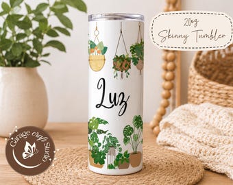 May include: White 20oz skinny tumbler featuring a botanical design with hanging plants and potted plants. The name "Luz" is written in a cursive font. The tumbler has a silver lid and is decorated with green and brown illustrations.