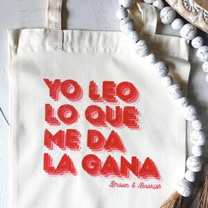 Könnte beinhalten: Cremefarbene Tragetasche mit rotem Text "YO LEO LO QUE ME DA LA GANA" und "Brown & Bookish". Die Tasche hat zwei Griffe. Eine dekorative Perlenkette und Quasten sind im Hintergrund. Der Text ist fett gedruckt.