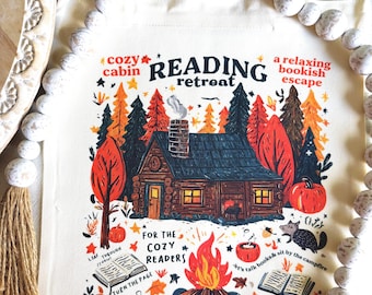 May include: Cream tote bag featuring a cabin scene with text: "Cozy Cabin Reading Retreat". The illustration includes a cabin, trees, a bonfire, and text such as "a relaxing bookish escape" and "Storytime by the fire".