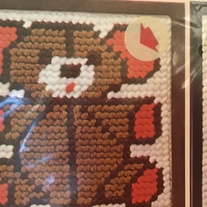 May include: A brown and orange teddy bear is stitched on a white canvas. The bear has a white face with a red nose. The canvas is framed in a brown frame.