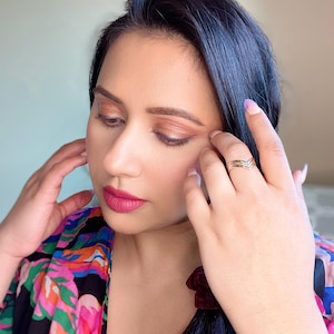 May include: A woman with dark hair and makeup, wearing a colorful floral patterned top. She has a gold ring on her finger and a burgundy scrunchie in her hair. Her lips are a bright pink color.