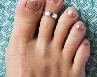 May include: A silver toe ring with a hammered finish. The ring is worn on the big toe of a foot with pink glitter nail polish.