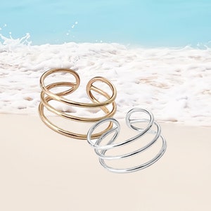 May include: Two minimalist ear cuffs, one gold and one silver, with a triple-ring design. The cuffs are shown against a beach background.