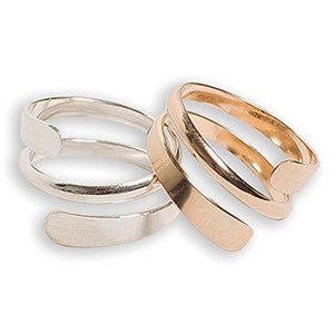 May include: Two spiral rings, one silver and one gold, are displayed. The rings have a minimalist design, with a flat, rectangular band that wraps around the finger. The gold ring has a warm, metallic hue, while the silver ring has a bright, polished finish.