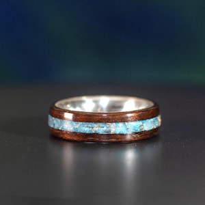 May include: A silver ring with a wooden band and a turquoise inlay. The ring has a polished silver interior and a dark brown wooden exterior. The turquoise inlay is a bright blue color and is set in the middle of the band.