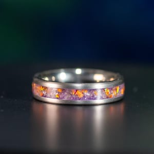 May include: A silver-toned metal ring featuring a central inlay of crushed purple and orange material. The band has a smooth, brushed finish on the outer edges, framing the vibrant, textured inlay that runs continuously around the circumference.