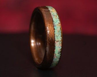 May include: A polished dark brown wooden ring featuring a central inlay of crushed light green stone fragments with subtle iridescent orange and yellow flecks embedded throughout the band, creating a textured appearance against the smooth wood.