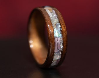 May include: A dark brown wooden ring featuring a central inlay of crushed iridescent shell fragments. The inlay displays a mix of white, light blue, and soft purple tones embedded within the polished wood band, creating a textured surface.