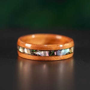 May include: A polished wooden ring featuring a central inlay of iridescent abalone shell fragments. The band has a smooth, rounded profile with a warm, light brown wood grain finish, contrasting with the shimmering, multi-colored shell pieces.
