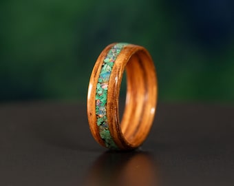 May include: A polished wooden ring featuring a central inlay of crushed green and iridescent stone fragments. The band displays a natural wood grain pattern with a smooth, glossy finish, showcasing a circular shape and a distinct, textured inlay.