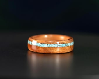 May include: A polished wooden ring featuring a horizontal inlay of small, light blue crushed stone fragments running through the center of the band. The wood has a warm, reddish-brown tone with a visible grain pattern and a smooth, glossy finish.