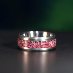 Op de afbeelding: Een zilveren ring met een band van roze en rode fragmenten. De ring heeft een gepolijste, metalen afwerking en een brede band gevuld met kleine, kleurrijke stukjes. De ring staat tegen een donkere achtergrond.