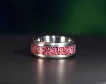 Puede incluir: Un anillo de metal plateado con una incrustación central de fragmentos de piedra triturada en tonos rosa y magenta. La banda tiene un acabado liso y pulido en los bordes superior e inferior, enmarcando la incrustación rosa texturizada y facetada.