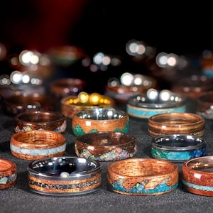 May include: A collection of wooden rings with various inlays, including turquoise, stone, and metal. The rings feature a variety of wood tones, from light to dark, and are displayed against a dark background. Some rings have a silver or gold band.