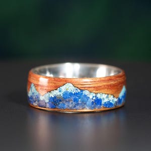 May include: A wooden ring with a blue and brown mountain scene inlay. The ring is made of wood and has a silver band.