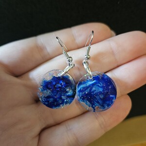 May include: A pair of round, blue resin earrings with silver-colored ear wires. The earrings have a swirling blue and white design with flecks of gold and silver. The earrings are held in a hand.