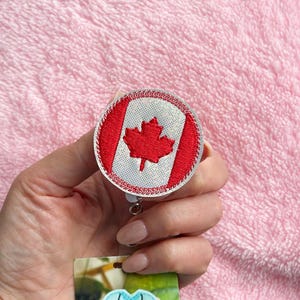 May include: A round retractable badge reel displaying the Canadian flag. The design features a red maple leaf on a white background, enclosed by red borders. The badge reel is held in a hand against a pink backdrop.