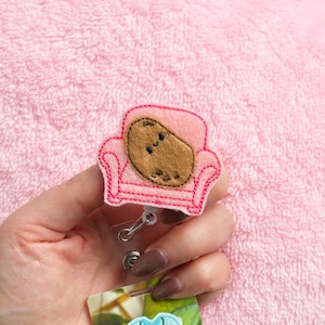 Könnte beinhalten: Ein pinkfarbenes Filz-Badge-Reel mit einem braunen Kartoffel-Charakter, der auf einer pinkfarbenen Couch liegt. Die Couch hat rote Nähte und ein detailliertes Design. Das Badge-Reel hat einen silbernen Clip und einen transparenten Kunststoffriemen.