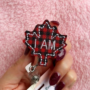 May include: A red and black plaid maple leaf badge reel with the text "I AM" in white. The badge reel has a white stitched outline and a retractable cord with a metal clip.