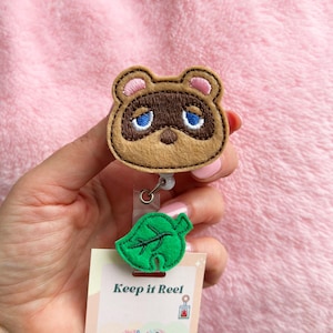 May include: A badge reel with a tan and brown cartoon bear face and blue eyes. Below the bear is a green leaf and a card that reads "Keep it Reel". The badge reel is attached to a silver retractable cord.