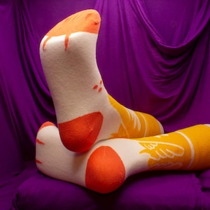 Könnte beinhalten: Ein Paar Socken mit weißer Basis, orangefarbenen Zehen und Fersen sowie orangefarbenen und gelben Akzenten. Die Socken haben ein Design eines Vogels oder Flügels an der Seite. Der Hintergrund ist ein tiefvioletter Stoff.