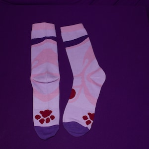 Puede incluir: Un par de calcetines rosa y morado con un diseño de huella de pata. Los calcetines tienen un diseño de mariposa rosa en la parte delantera y una huella de pata roja en el talón.