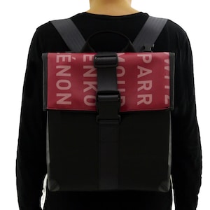 Può includere: Uno zaino nero con una patta rossa che presenta il testo "PARR" e "ENON" in rosa. Lo zaino ha una fibbia nera e spalline regolabili.