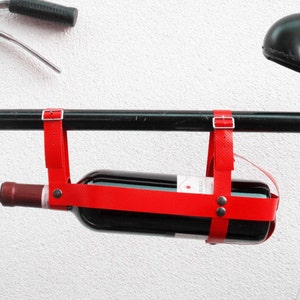 Può includere: Un porta bottiglie di vino in pelle rossa attaccato al manubrio di una bicicletta. Il porta bottiglie ha due cinghie con fibbie e una bottiglia di vino rosso è fissata all'interno.