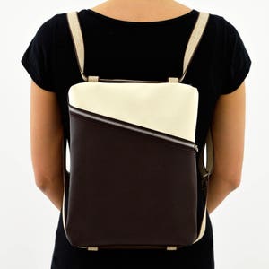 Può includere: Uno zaino in pelle marrone e crema con una cerniera diagonale. Lo zaino ha un design semplice ed è indossato da una persona.