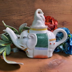 May include: White ceramic teapot shaped like an elephant with a small elephant figurine on top. The teapot has a green, blue, and orange design.