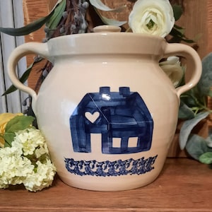 May include: A cream-colored ceramic crock with a blue and white house design. The house has a white heart on the front and a blue roof. The crock has two handles and a lid.