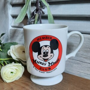 以下が含まれることがあります： 赤い円とミッキーマウスの顔が描かれた白いセラミック製のマグカップ。円には「Member Mickey Mouse Club」と「Walt Disney Productions」の文字が書かれています。