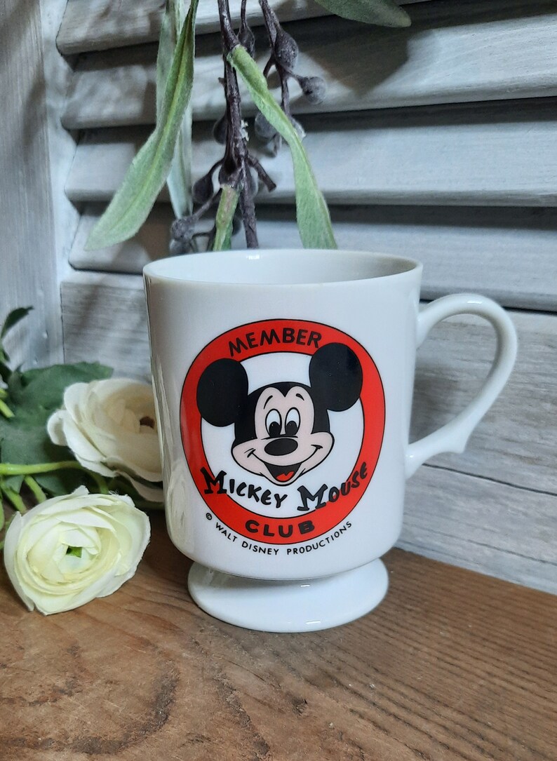 以下が含まれることがあります： 赤い円とミッキーマウスの顔が描かれた白いセラミック製のマグカップ。円には「Member Mickey Mouse Club」と「Walt Disney Productions」の文字が書かれています。