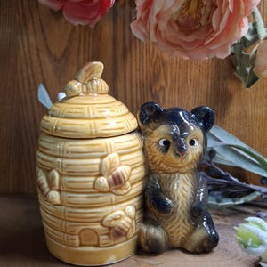 May include: A ceramic honey pot shaped like a beehive with a bear figurine next to it. The beehive has a lid with a bee on top and the bear is brown with black accents.