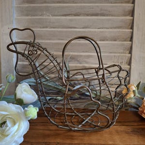 May include: A decorative wire chicken basket with a handle. The basket is made of bronze-colored metal and has a handle. The chicken basket is sitting on a wooden surface with flowers in the foreground and a shuttered background.