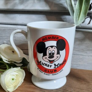 以下が含まれることがあります： 白いセラミック製のマグカップで、赤い円の中にミッキーマウスの顔が描かれています。円には「メンバー ミッキーマウス クラブ &copy; ウォルト・ディズニー・プロダクションズ」と書いてあります。