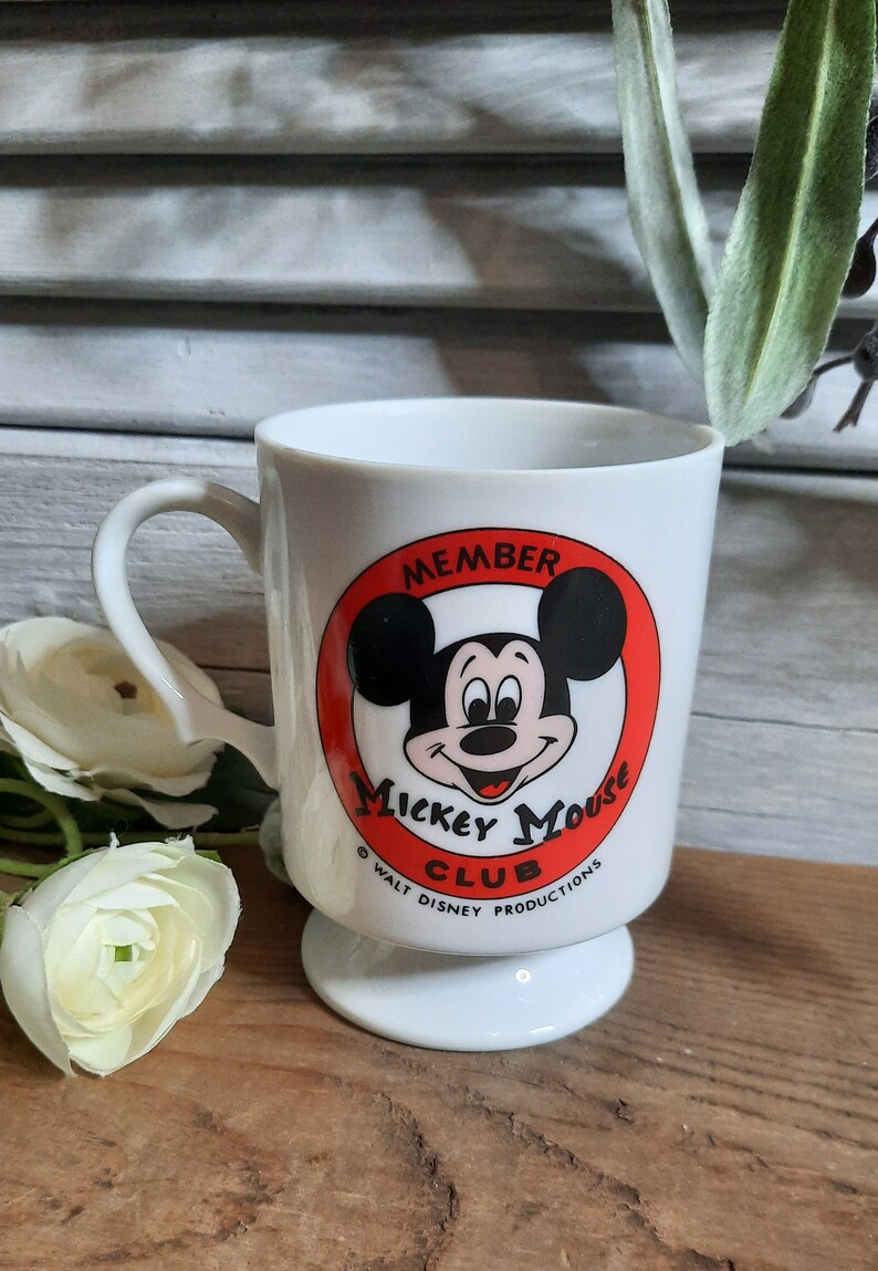 以下が含まれることがあります： 白いセラミック製のマグカップで、赤い円の中にミッキーマウスの顔が描かれています。円には「メンバー ミッキーマウス クラブ &copy; ウォルト・ディズニー・プロダクションズ」と書いてあります。