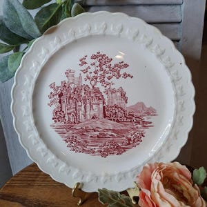 May include: White ceramic plate with a scalloped edge and a red and white landscape design. The plate features a detailed illustration of a castle, trees, and a mountain range. The plate is displayed on a wooden surface.