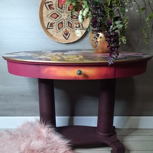 May include: A burgundy oval table with a single drawer and a decorative gold knob. The table top features a colorful, abstract design. The table has two turned legs and a lower shelf.
