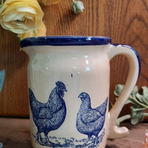 May include: A white ceramic pitcher with a blue rim and handle. The pitcher features a blue and white design of two chickens.