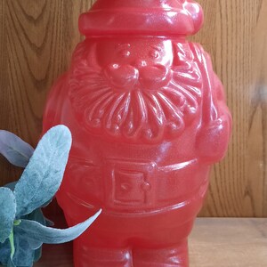 May include: A red, sparkly Santa Claus figurine with a white beard and a white hat. The figurine is made of plastic and has a shiny, glittery finish.