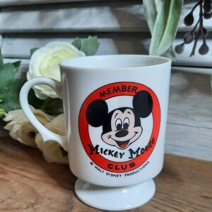 以下が含まれることがあります： 赤い円とミッキーマウスの顔が描かれた白いセラミック製のマグカップ。円には「Member Mickey Mouse Club」と「Walt Disney Productions」の文字が書かれています。