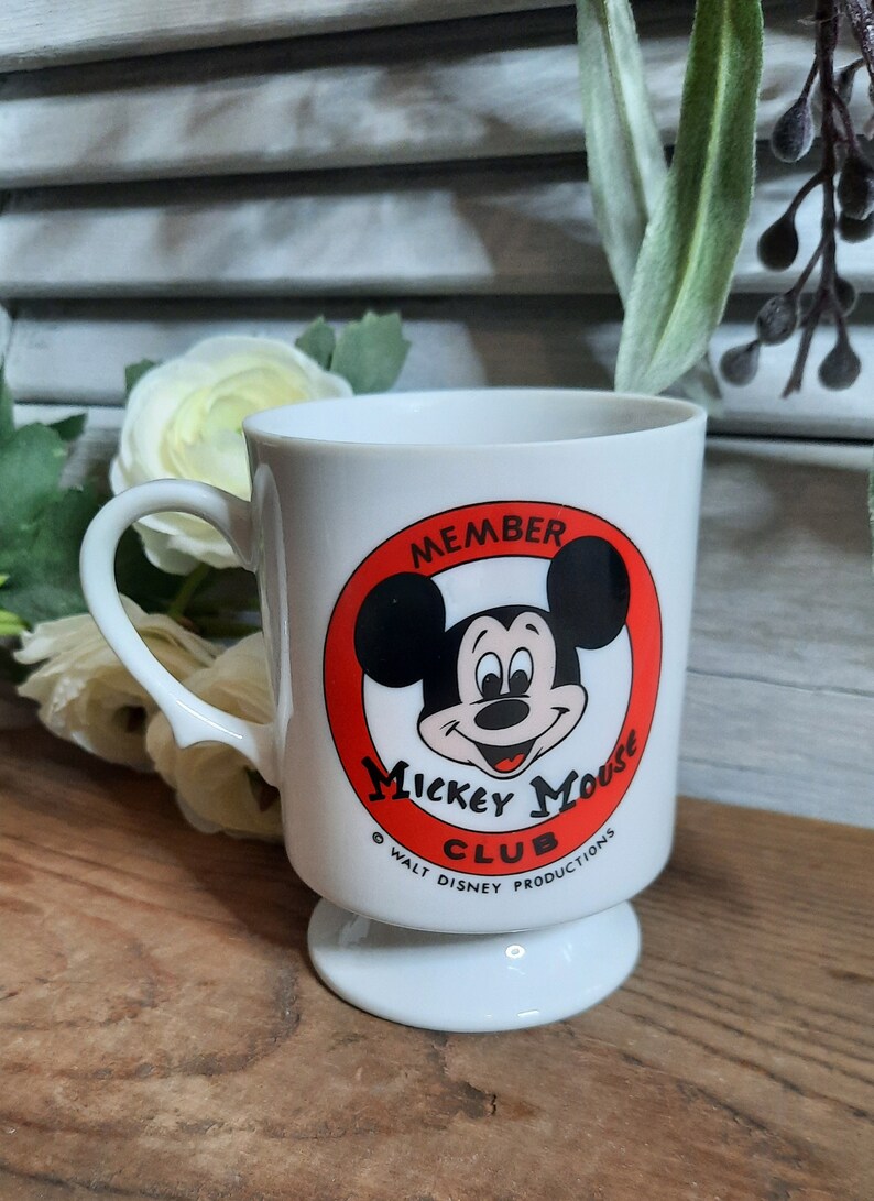 以下が含まれることがあります： 赤い円とミッキーマウスの顔が描かれた白いセラミック製のマグカップ。円には「Member Mickey Mouse Club」と「Walt Disney Productions」の文字が書かれています。