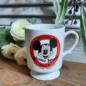 以下が含まれることがあります： 赤い円とミッキーマウスの顔が描かれた白いセラミック製のマグカップ。円には「Member Mickey Mouse Club &copy; Walt Disney Productions」と書かれています。