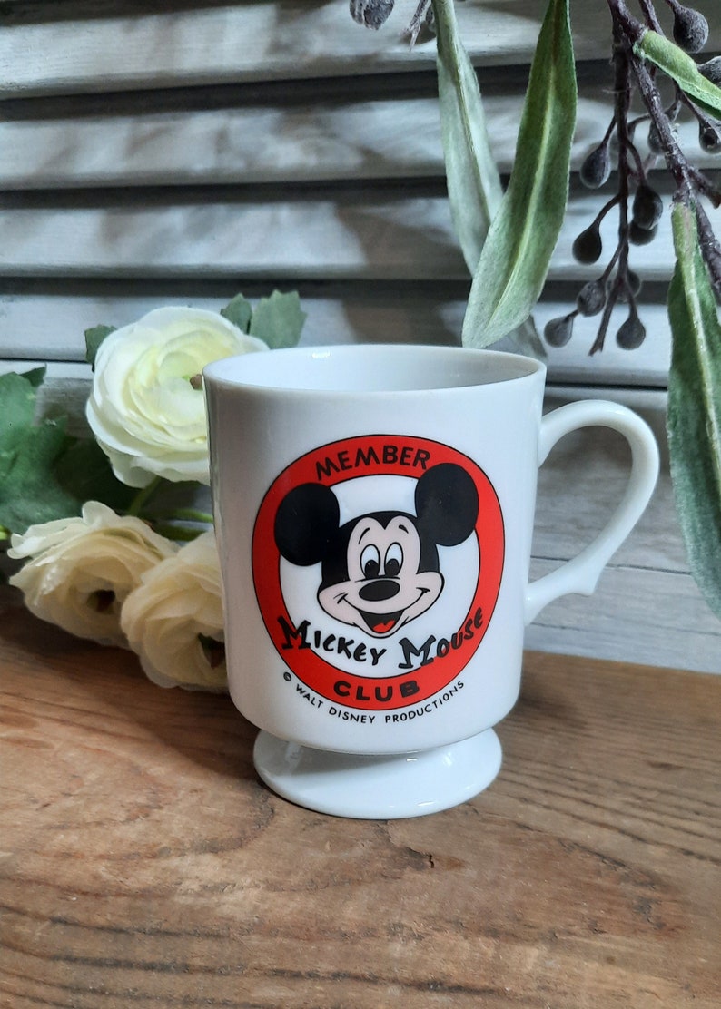 以下が含まれることがあります： 赤い円とミッキーマウスの顔が描かれた白いセラミック製のマグカップ。円には「Member Mickey Mouse Club &copy; Walt Disney Productions」と書かれています。