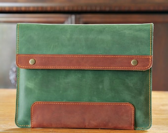 Puede incluir: Funda para portátil de cuero verde con detalles en cuero marrón y una solapa asegurada con dos remaches de latón. La funda rectangular está sobre una superficie de madera. Las costuras son visibles.