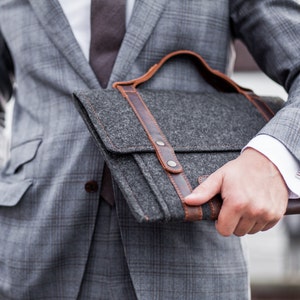 May include: A gray felt briefcase with brown leather straps and a metal clasp. The briefcase is held by a person wearing a gray suit.