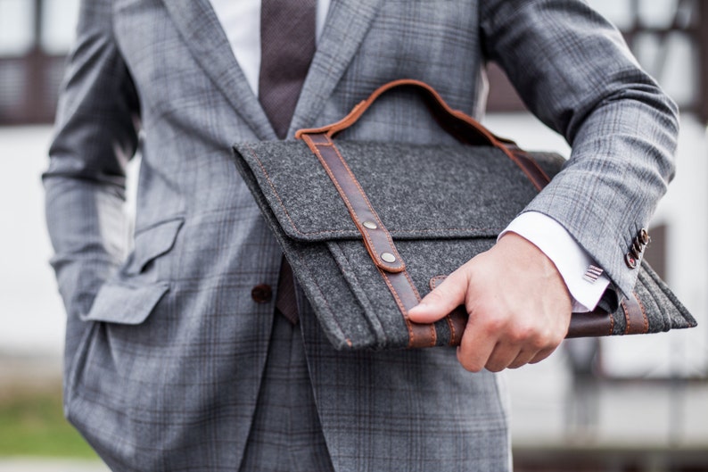 May include: A gray felt briefcase with brown leather straps and a metal clasp. The briefcase is held by a person wearing a gray suit.