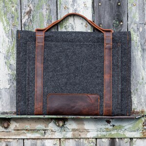 May include: A gray felt laptop sleeve with brown leather accents and a top handle. The sleeve is sitting on a weathered wooden surface.
